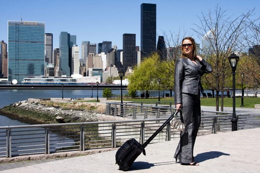 Beautiful Caucasian Latina entrepreneur business woman in grey suit and sunglasses with a carry-on luggage and purse standing in a park in New York City in front of the skyline and the United Nations buildings.