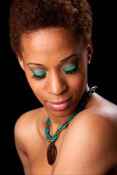 Beautiful face of an African American female fashion model looking down showing her eyelids with green eyeshadow and long eyelashes, bare shoulders, and wearing a green with brown necklace, isolated.