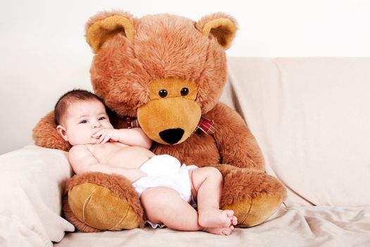 Cute Caucasian Hispanic unisex baby in arms of a big brown stuffed teddy bear sitting on couch.