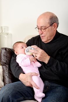 Caucasian Hispanic Grandfather feeding cute baby girl with a bottle of milk while sitting on a couch in a livingroom holding infant in his arms.