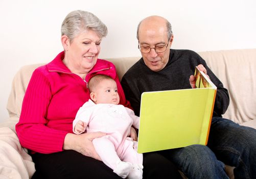 Happy fun Caucasian Hispanic Middle Eastern family sitting together on couch reading book to baby. Grandfather and Grandmother reading green book to infant granddaughter.