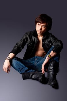 Handsome Asian male wearing leather jacket over a bare chest and jeans with macho attitude while sitting on floor expressing thought, isolated.