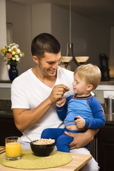 Caucasian man feeding toddler son on lap in kitchen.