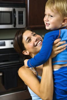 Caucasian woman in kitchen lifting smiling toddler son into the air.
