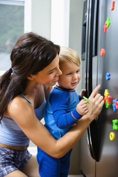 Caucasian toddler boy and mother playing with magnets on refrigerator.