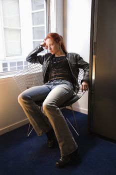 Pretty redhead young woman sitting indoors on metal chair by window looking out.
