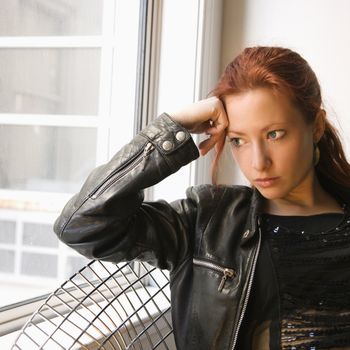Pretty redhead young woman sitting indoors on metal chair looking out window.