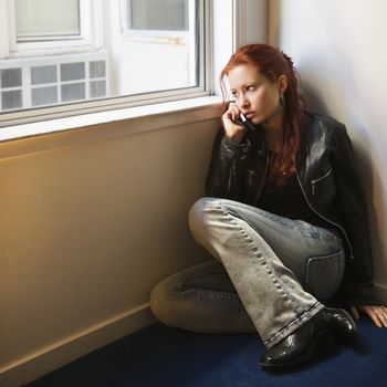 Pretty redhead young woman sitting on floor indoors looking out window holding cellphone to ear.