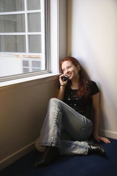 Pretty redhead young woman sitting on floor indoors by window holding cellphone to ear smiling.