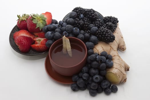 Ginger tea in a handleless cup surrounded by strawberries, blueberries, blackberries, and ginger root