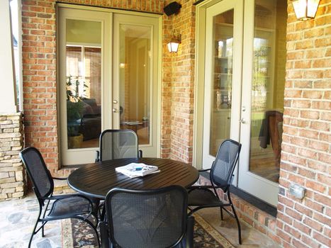 A wood and metal table and chairs on a covered stone patio on an american brick home. Two large glass french doors open up to the house in the background