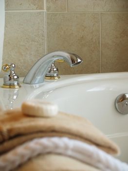 Faucet and handles on the side of a bathtub with towels and a bar of soap blurred in the foreground