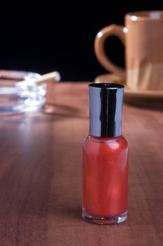 Red nail polish standing on a coffee table in front of a cup of coffee and an ashtray.