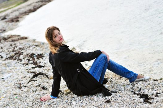 pensive  teen girl sitting on the pebble close to sea in spring time
