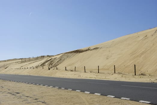 An isolated road along big dunes with a little barrier and some stranding in the sand. And the sky is perfectly blue totally without cloud