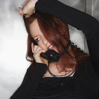 Portrait of pretty young redhead woman holding telephone receiver to ear with cord wrapped around head smiling.