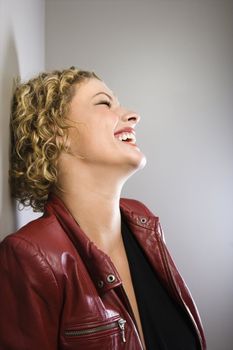 Profile of Caucasian young adult woman in red jacket leaning back against wall laughing.
