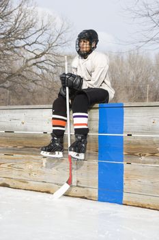 Boy in ice hockey uniform holding hockey stick sitting on sidelines.