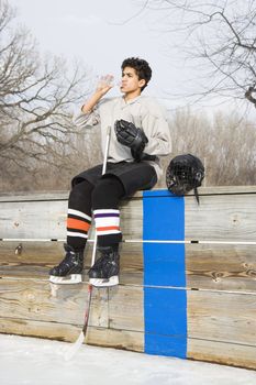 Boy in ice hockey uniform holding hockey stick sitting on sidelines drinking water.