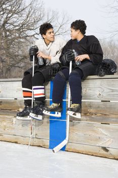 Two boys in ice hockey uniforms sitting on ice rink sidelines talking to eachother.