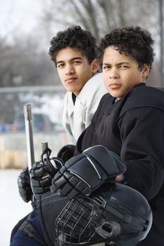 Two boys in ice hockey uniforms sitting on ice rink sidelines looking.