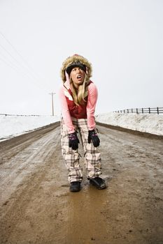 Young woman in winter clothes standing alone in middle of muddy dirt road and snow making strange facial expression.