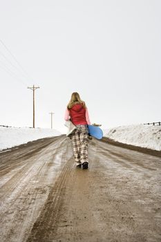 Young woman in winter clothes walking alone down muddy dirt road carrying snowboard and boots.