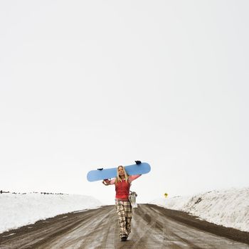 Young woman in winter clothes walking alone down muddy dirt road carrying snowboard and boots.