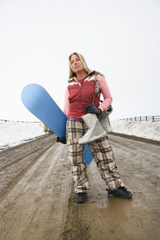 Young woman in winter clothes standing alone on muddy dirt road holding snowboard and boots.