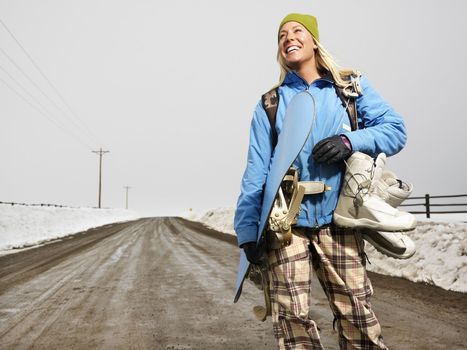 Young woman in winter clothes standing on muddy dirt road holding snowboard and boots smiling.