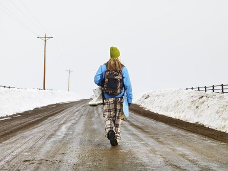 Young woman in winter clothes standing on muddy dirt road holding snowboard and boots.