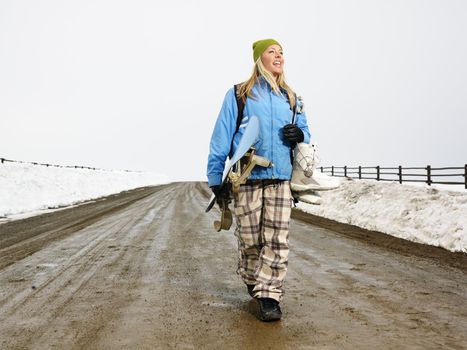 Young woman in winter clothes walking down muddy dirt road holding snowboard and boots smiling.