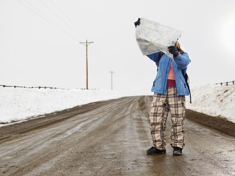 Young woman in winter clothes standing on muddy dirt road looking at map.