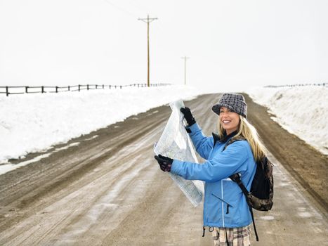 Young woman in winter clothes standing on muddy dirt road holding open map smiling.