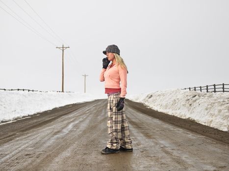 Young woman in winter clothes standing on muddy dirt road talking on cell phone.