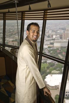 Portrait of young adult male Indian standing near window in tower restaurant smiling at viewer.