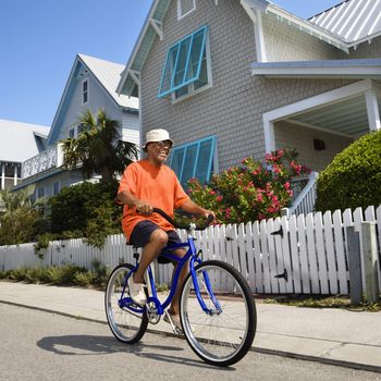 Middle aged African American man bicycling down street next to homes and fence.