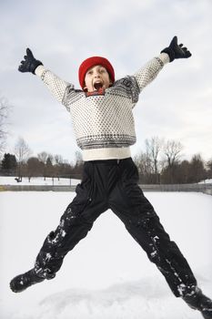 Excited Caucasian boy wearing sweater and red winter cap jumping in air with arms and legs outstretched.