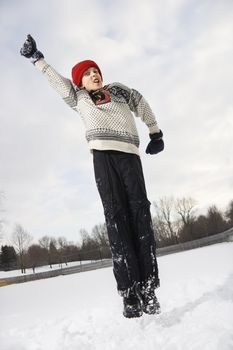 Caucasian boy wearing sweater and red winter cap jumping in air with arm outstretched.