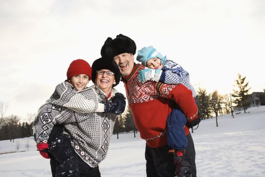 Caucasian middle aged parents carrying their Caucasian son and daughter piggyback style smiling at viewer.