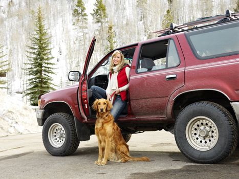 Woman with dog sitting with dirt splattered SUV automobile in snowy rural setting.