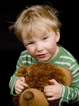 Smiling little boy with brown teddy-bear on black background. Boy have blue eyes, blond hair and a bit of dirt on his face