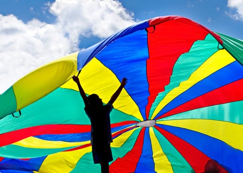 silhouette of child in play backlit with a colorful parachute floating in the breeze with blue sky and clouds above