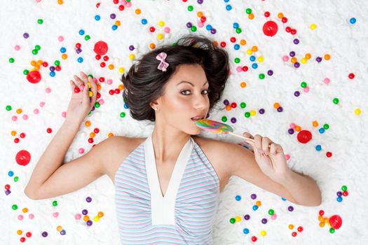 Young Latina woman laying on ruffled cloud like floor between colorful bubblegum balls eating a lollipop