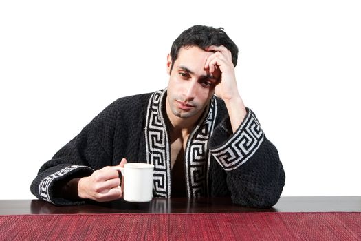 Handsome guy in the morning who just woke up sitting at a table in his robe with a cup recovering from his hangover, isolated on white