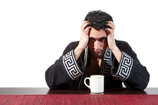 Handsome guy in the morning who just woke up sitting at a table in his robe and hands in his hair with a cup, isolated on white