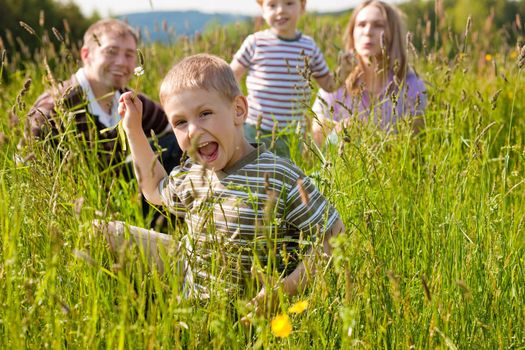 Very happy family with two kids sitting in a meadow in the summer sun in front of a forest and hills, they are nearly hidden by the high grass, very peaceful scene