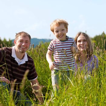 Very happy family with son sitting in a meadow in the summer sun in front of a forest and hills, they are nearly hidden by the high grass, very peaceful scene