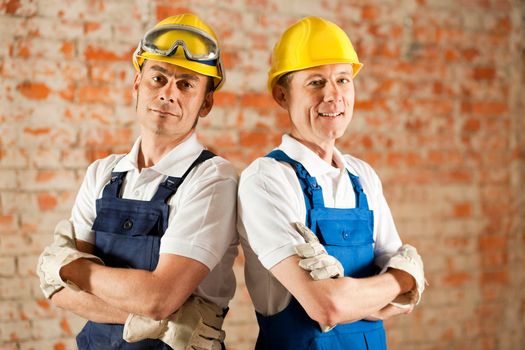 Two construction workers standing in a construction site in front of a war brick wall. Their arms are folded and they are wearing hardhats
