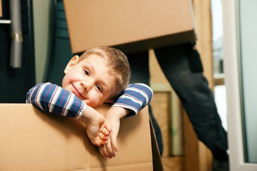 Family moving in their new home. The son is sitting inside a moving box. In the background the father - or a mover (only legs to be seen) is carrying boxes inside the building